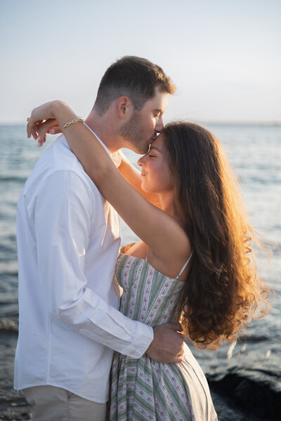 Rhode Island Wedding Photographer | Kelsey Sheehan Photography Timeless Rhode Island Weddings | A couple embraces by the ocean at sunset. The man in a white shirt kisses the woman's forehead as she smiles, creating a romantic and peaceful tone.