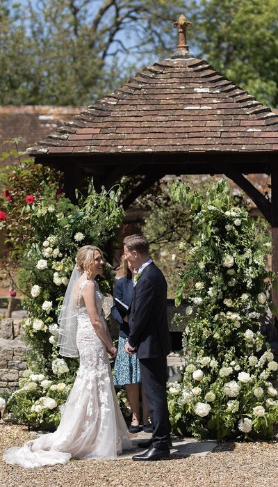 Bride and Groom exchanging vows under outdoor wedding pergola at Eden Barn Cumbria Lake District with flower arch flower installation apricot flowers pampas eucalyptus
