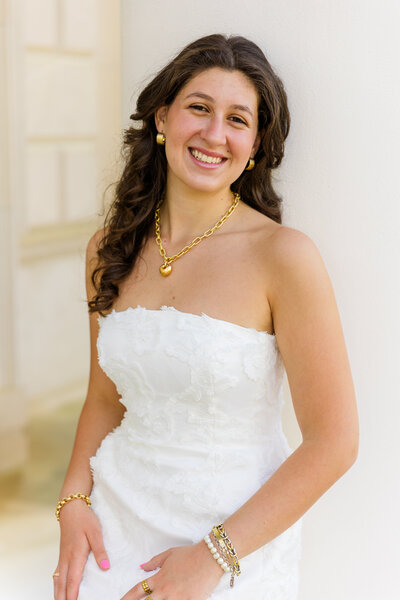 top body frame of a senior girl laughing at the camera leaning against a pole wearing a white dress standing in front of a clear color solid wall during graduation photoshoot with Atlanta photographer Laure Photography