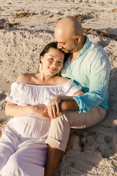 Maternity couple sitting on the sand in Sunny Isles Beach, with the expecting mother smiling and leaning into her partner.