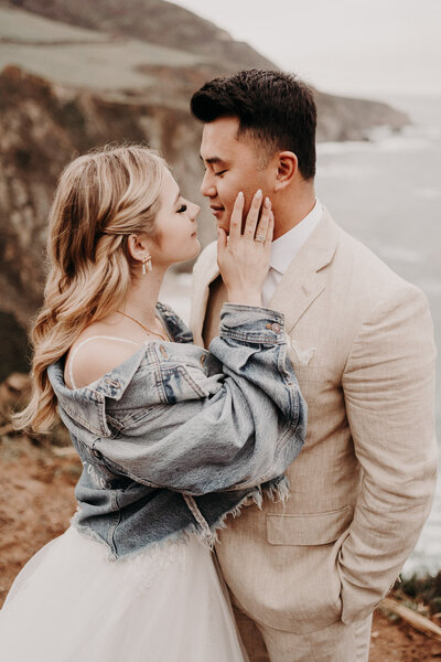 a bride stroking a groom's face while standing on a cliff side