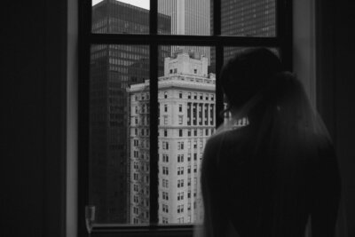 Black and white photo of silhouette of bride wearing a veil looking through a window with a view of Chicago skyscrapers.