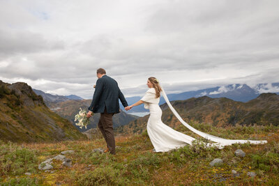 A bride and groom walk along a remote Alaskan beach toward a waiting helicopter, surrounded by rugged coastline and dense evergreen forests. The groom carries a backpack, and the bride’s wedding dress flows behind her, capturing the essence of an adventurous elopement in the Alaskan wilderness.