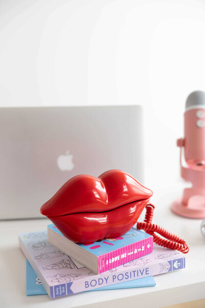 A stack of Emily's books (Body Positive and I Love Me from A to Z) with a lip shaped phone sitting on top and her laptop and podcast microphone in the background.