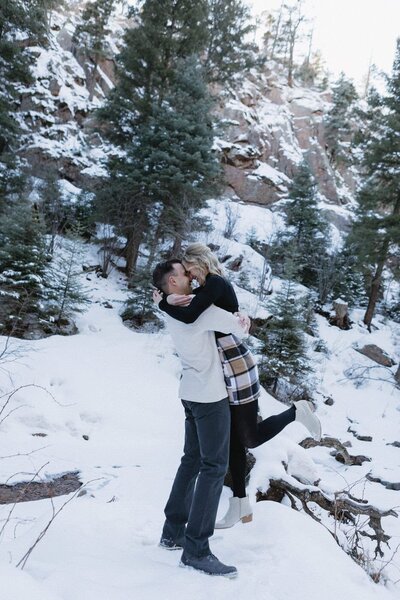 A couple stands in a snowy landscape with pine trees and he lifts her off the ground.