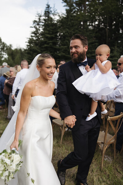 A couple in formal attire, with the woman in a white gown and the man in a black suit, seated on an outdoor stone bench, both smiling; lush greenery and flowers form the background, perfectly captured by a Canadian wedding planner.