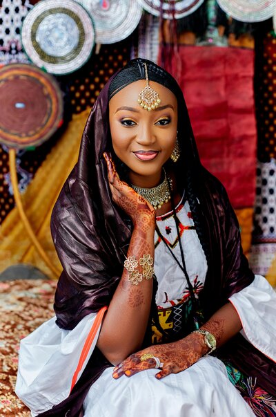 Elegant Nigerian bride wearing traditional attire with intricate henna, gold jewellery, and a rich headscarf, smiling in front of a vibrant cultural backdrop