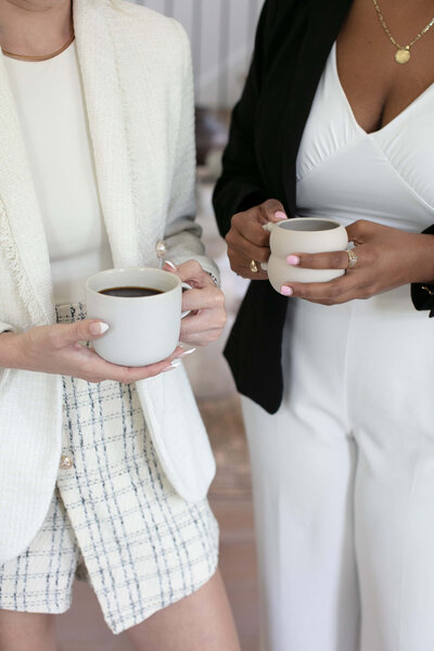 Two women stand next to each other, holding coffee cups and engaging in conversation.