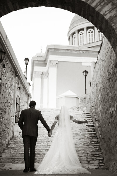 Bride and groom walking hand in hand up a stone staircase in Greece, with a grand church and classical architecture in the background.