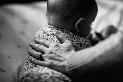 Black and white image of a mother's hand rubbing her 4 month old baby's back. Baby is alert on her tummy in her crib: we just see the back of her head looking up, and the floral print of her onesie.