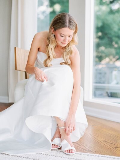 Rhode Island Wedding Photographers | A bride in a white gown sits on a chair, adjusting her shoe. Soft natural light streams in through large windows.