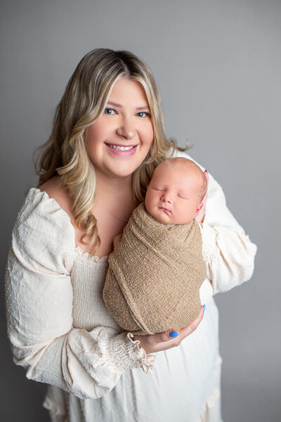 Blonde mother holding baby boy wrapped in beige color for a simple modern newborn portraiture in studio 