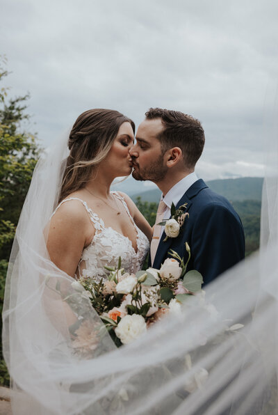 Knoxville wedding photographers photographs bridal portrait of bride and groom kissing in the mountains as the brides veil flows in front of the couple