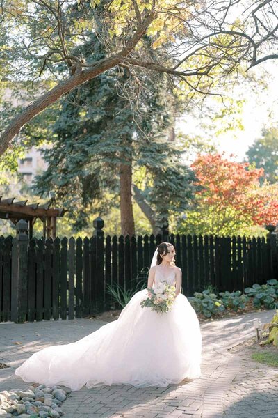 Bride in voluminous gown sitting in sunlight with bouquet at Old Mill garden, romantic wedding photography in Toronto