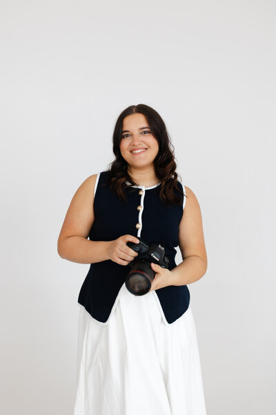 Wedding photographer smiles while holding camera against white backdrop wearing navy and white outfit