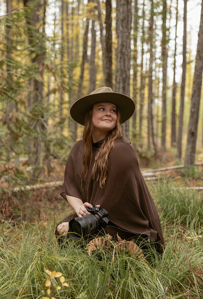Photographer sitting in the forest with camera in hand, wearing a brown poncho and green hat surrounded by tall trees.