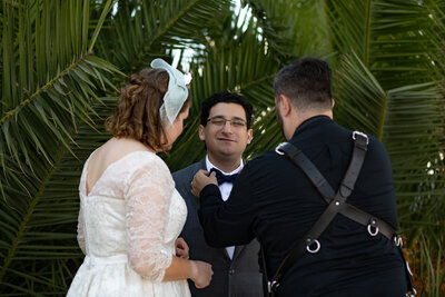 A photographer adjusting a groom's bow tie as they stand next to the bride.