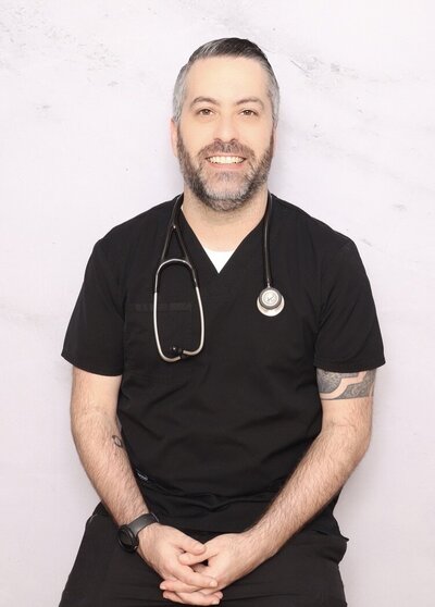 Male doctor wearing black scrubs and a stethoscope, smiling in front of a light-colored photo booth backdrop, ideal for professional headshots.