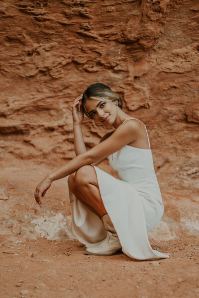 A bride wearing a casual white dress and white cowgirl boots, almost sitting on the red rocks but not quite. Touching her hair with one hand with the other balanced on her knee.
