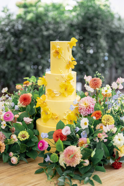 A three-tiered yellow cake decorated with yellow flowers, surrounded by vibrant floral arrangements featuring pink, orange, and blue blossoms, set against a lush green background.