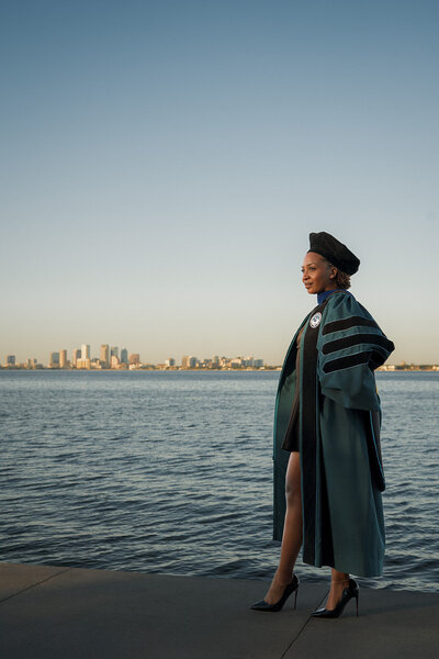 woman stands proudly with doctoral cap and gown at sunset with Tampa skyline in the background
