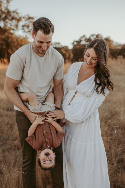 An expecting mother in a pretty white gown laughs with her husband who is holding their toddler boy upside down in a El Dorado Hills, CA field.  