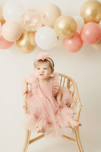 1 year old sitting on a chair on a white backdrop with balloons in the background