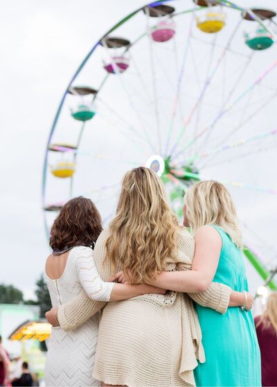 three girls side hugging each other as they stand and look at a Ferris wheel at carnival in Hampton Roads Virginia with best family photographer near me