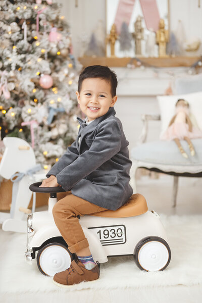 Little boy sitting on a vintage toy car smiling wearing a velvet holiday suite inside a nutcracker themed studio set in Phoenix