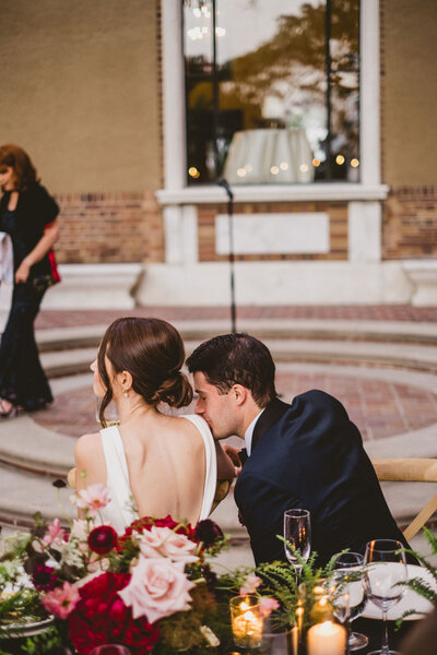 A photo of a bride and groom from behind while they sit at their reception table. The groom is kissing the brides shoulder.