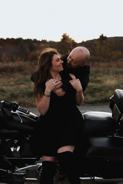 Adventurous couple sitting on motorcycle shares warm embrace in all black dress and black jean outfits