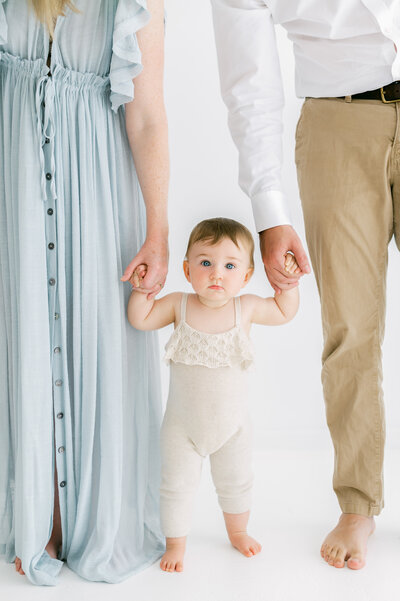 Baby girl looking at camera with neutral expression while mum and dad hold each of her hands