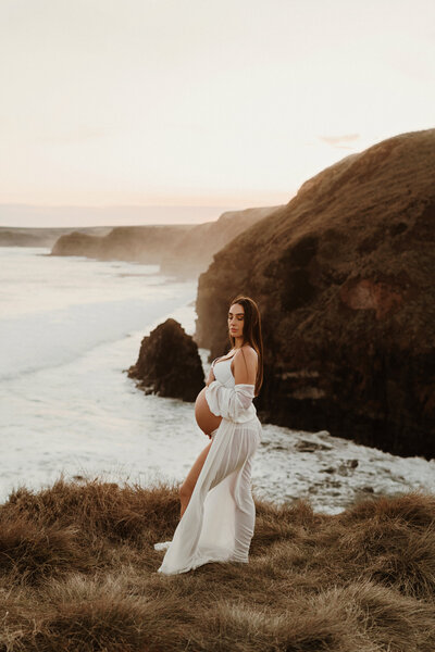 Pregnancy photoshoot of a mum standing on a cliff with her arms up above her head. In a white crochet dress where her pregnant belly is visible through. Cliffs and water behind her in the background. Mornington Peninsula location