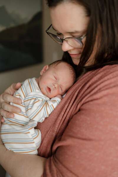 Mom holding and looking down lovingly at newborn baby in striped sleeper during in home newborn session with Alexis Adkins Photography in Firestone Colorado