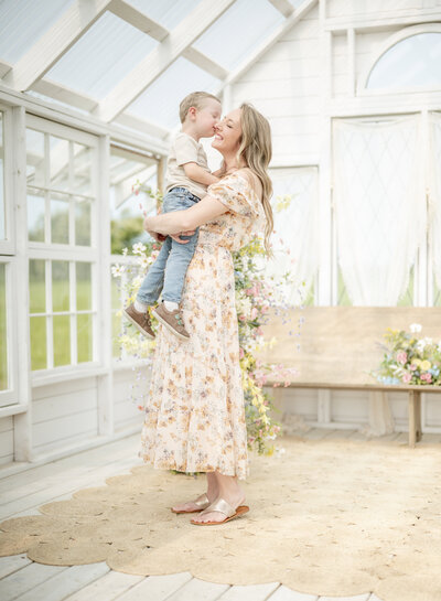 Joyful mom holding her son inside a bright, white greenhouse studio during a warm, light-filled Kansas City family photography session.