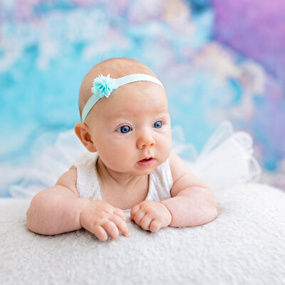 Baby girl with bright blue eyes lying on her tummy, wearing a light blue headband with a flower, against a soft pastel blue and purple backdrop.
