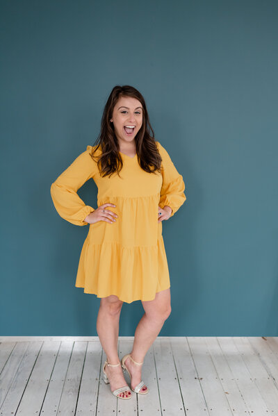 Bee, founder and owner of BusyBSocial, stares and smiles with her mouth wide with her hands on her hips wearing a yellow dress in front of a blue background