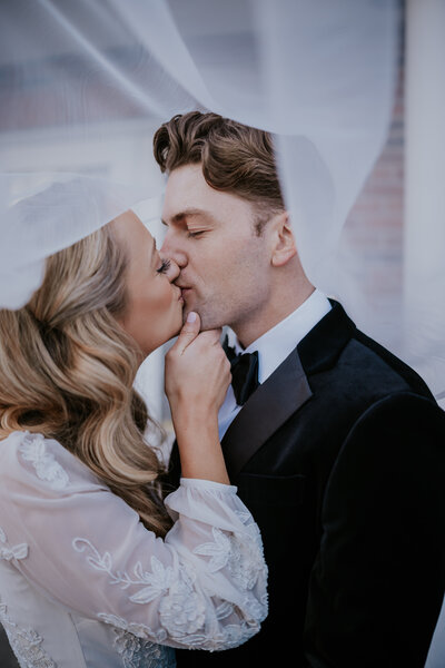 Bride and groom share a romantic kiss under the veil, with delicate lace details and soft lighting adding elegance to their wedding day.