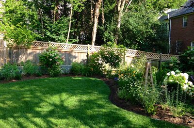 Transformed backyard featuring a lush green lawn, vibrant flower beds, a wooden privacy fence with lattice details, and bright, open landscaping.