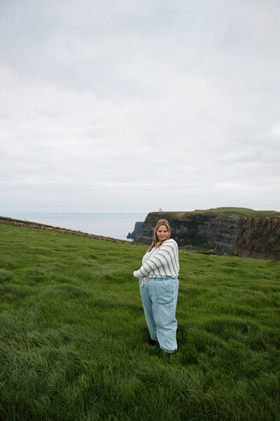 Photo of Hailey Lunsford of Hailey Lunsford Photography standing across from the Cliffs of Moher in Ireland. Hailey is wearing light blue jean joggers and a striped white and green sweater.