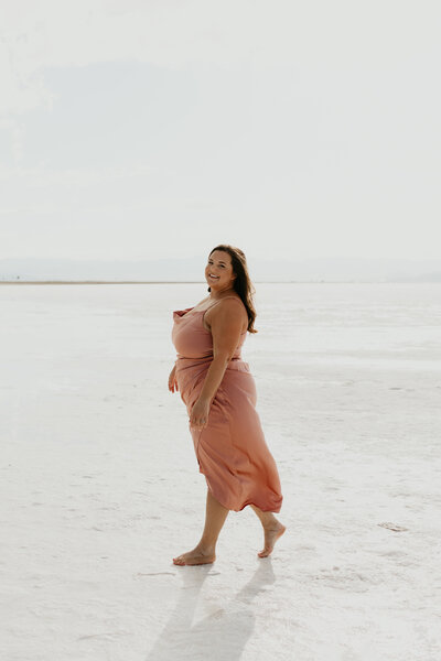 Refined editorial portrait of a woman walking barefoot in a pink silk dress at the Texas salt flats, evoking movement, femininity, and natural beauty.