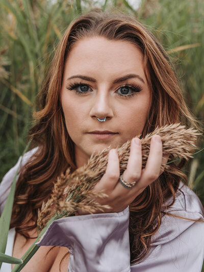 headshot portrait of a woman with long reddish-brown hair and blue eyes in a tall grassy field wearing a lavender, satin, long-sleeve blouse gently holding a fuzzy seed head from top of the grass located at Rocky Point State Park in Warwick, Rhode Island