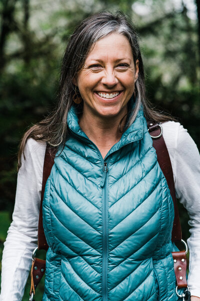 An elopement photographer wearing two Canon cameras and a blue puff vest smiles at the camera in the lush green PNW forest. | Erica Swantek Photography