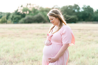 A pregnant mother dressed in a long pink dress stands in a field of tall grass at Brushy Creek Lake Park. She looks down at her belly and smiles, beautifully captured by an Austin Texas Portrait Photographer