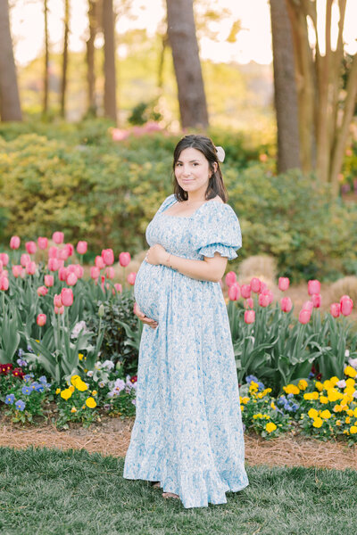 A glowing expectant mother in a blue floral dress standing among blooming tulips at golden hour — Portrait photography in Raleigh.