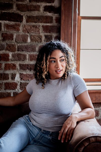 A woman sits on a brown leather couch with a brick wall behind her. She has curly hair and is looking out a window to her left. 