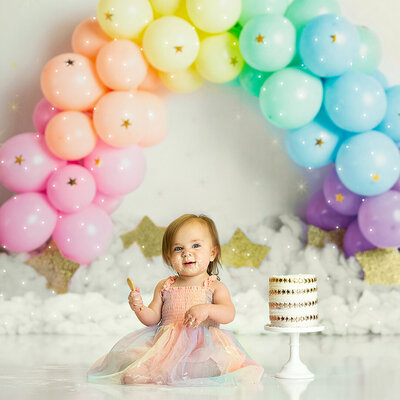 A baby girl is holding a spoon, sitting next to a cake, posed in front of a rainbow balloon backdrop in Warwick NY