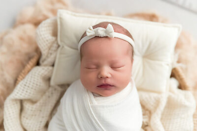 Baby girl sleeps on a pillow with a bow on her head and wrapped in white.