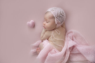 newborn baby girl in soft pink knit bonnet with draped pink wrap and felt heart