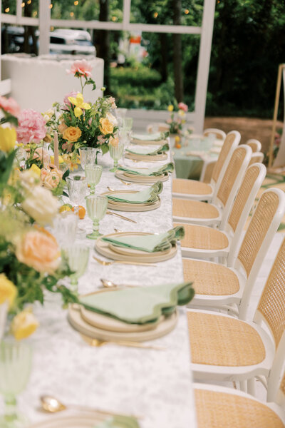 Side view of a long reception table styled with pastel florals, green napkins, and white cane-back chairs in a pavilion.